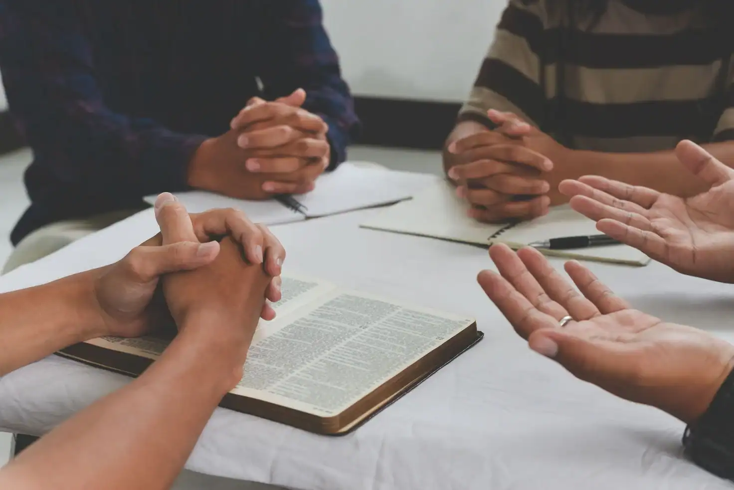 Believers gathered around a table with hands folded in prayer beside an open Bible